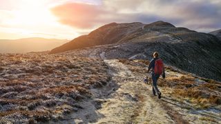 A woman with a red backpack walking along a hiking path towards the mountain summit of High Spy from Maiden Moor in the Lake District at sunrise