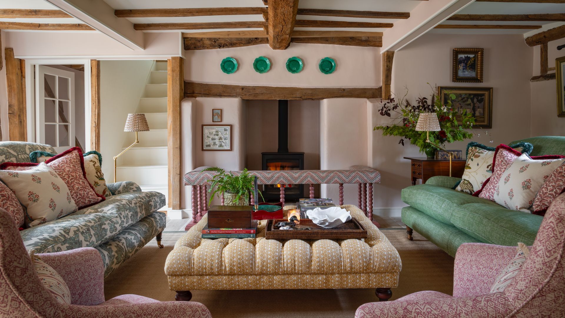 A country living room with a wood burning stove, two sofas opposite one another and two pink patterned armchairs in the foreground. In the middle of the room is a large yellow upholstered ottoman with a tray on top filled with books and plants.