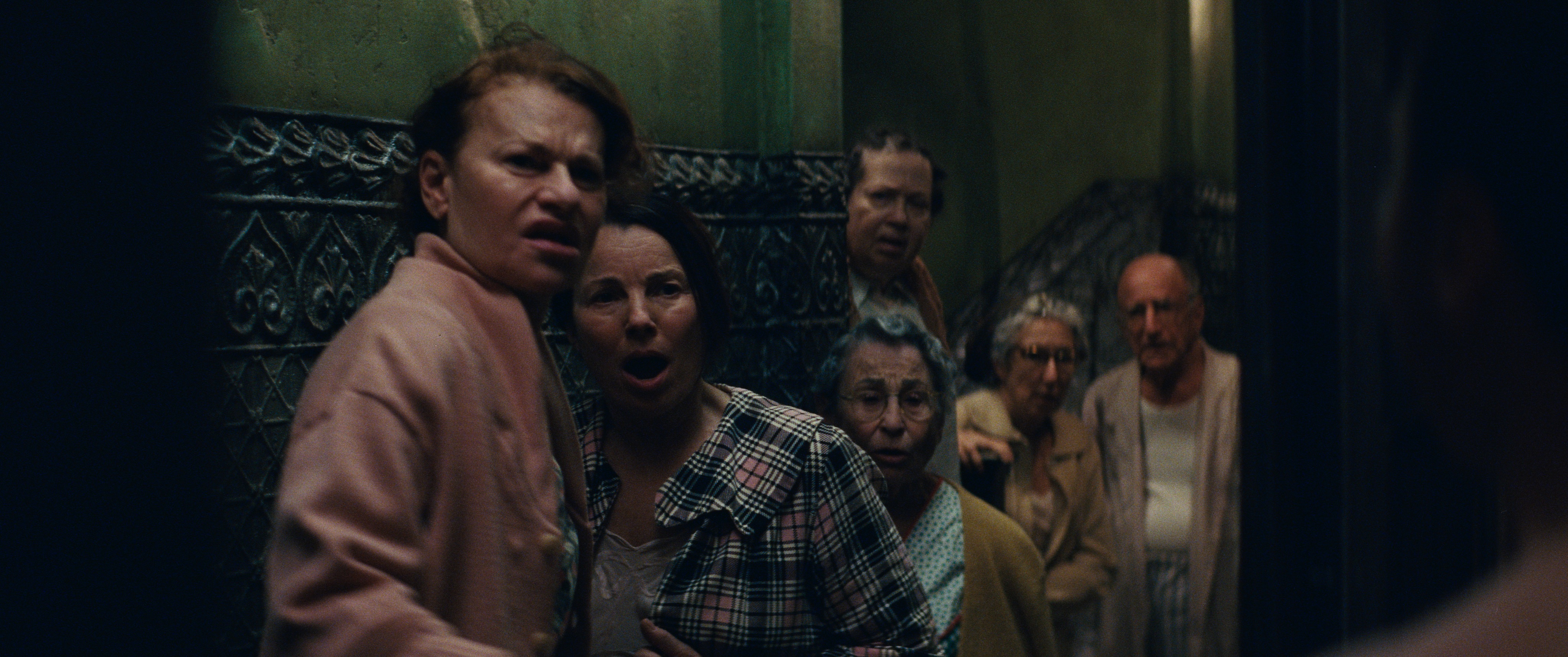 sandra bernhard and fran drescher as marty's family with a group of elderly neighbors standing in an apartment hallway looking shocked in the movie marty supreme