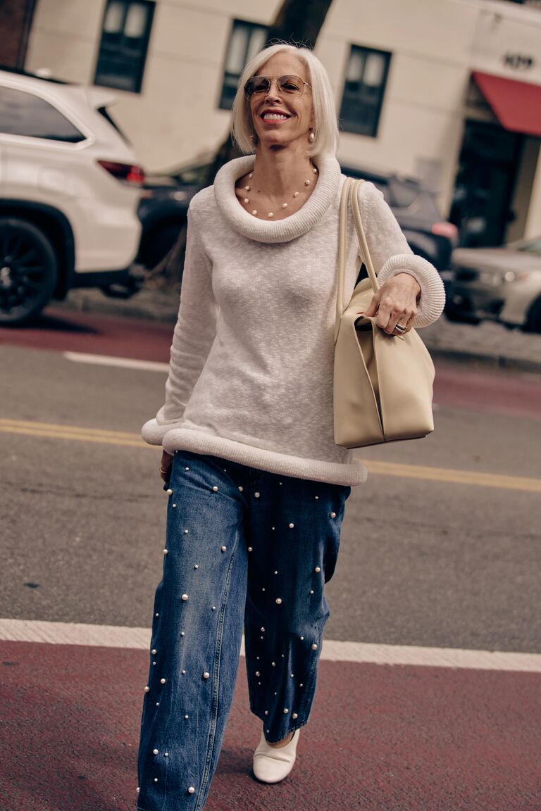 a woman at new york fashion week wearing a white round neck sweater and pearl embellished blue jeans