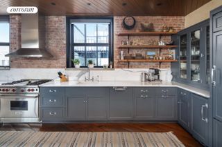 An open plan kitchen with gray-blue cabinets in an industrial style apartment with exposed brickwork and concrete columns