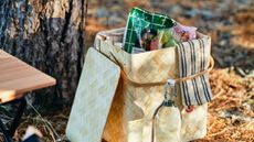 A woven wooden basket with a cooler bag full of snacks inside, on a floor with fallen pine needles.