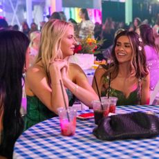 the women from secret lives of mormon wives sit around a table with a checkered table cloth wearing flower crowns and drinking dirty sodas in a still from mormon wives season 4