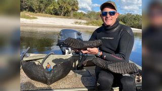 Man sits beside a river in a wetsuit holding a fossil engraved with lines, sitting next to two others found.