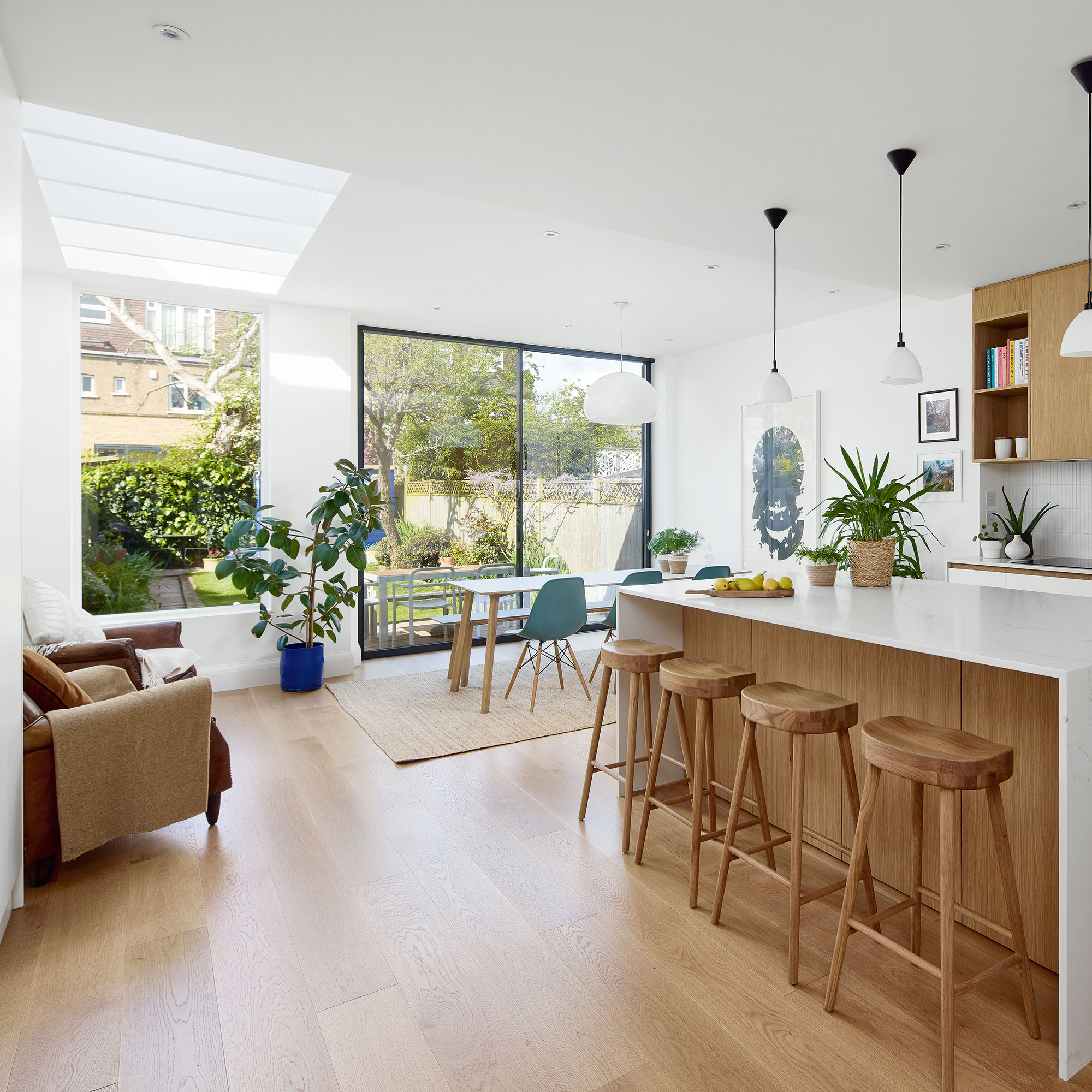 White kitchen with wooden floor, sleek cabinetry and white island with wooden bar stools. Skylights in roof and floor-to-ceiling windows out to garden