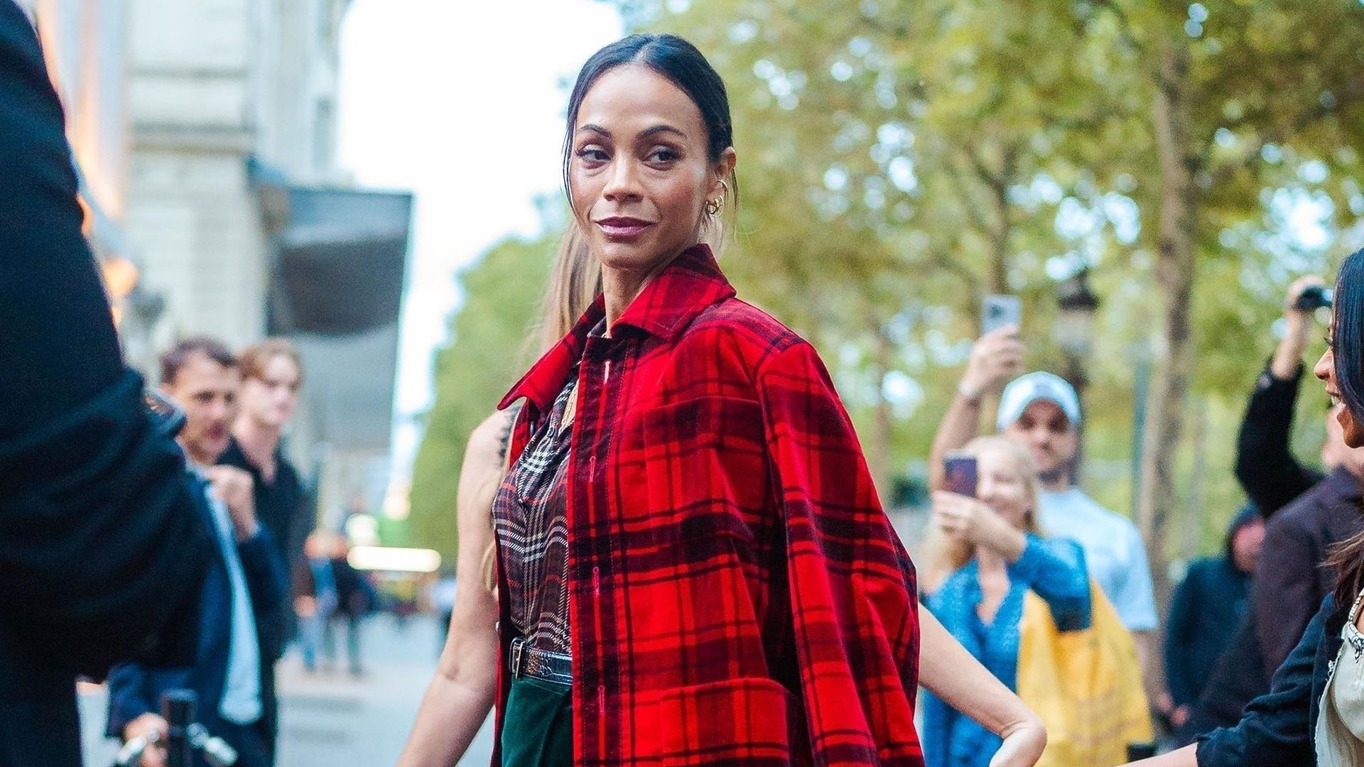 Zoe Saldana wearing a red plaid coat, plaid button down, velvet green skirt, and black leather knee-high boots
