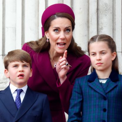 Kate Middleton looks stern while wearing a deep red coat dress and matching hat as she instructs children Prince George, Prince Louis, and Princess Charlotte