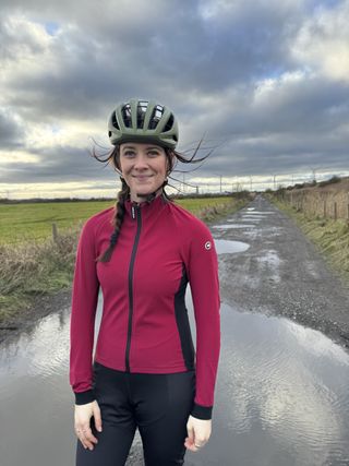 Woman faces the camera in pink jacket and green helmet in front of a muddy puddle.
