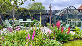 Garden with flowers at the front, a seating area to the left and a green house to the right