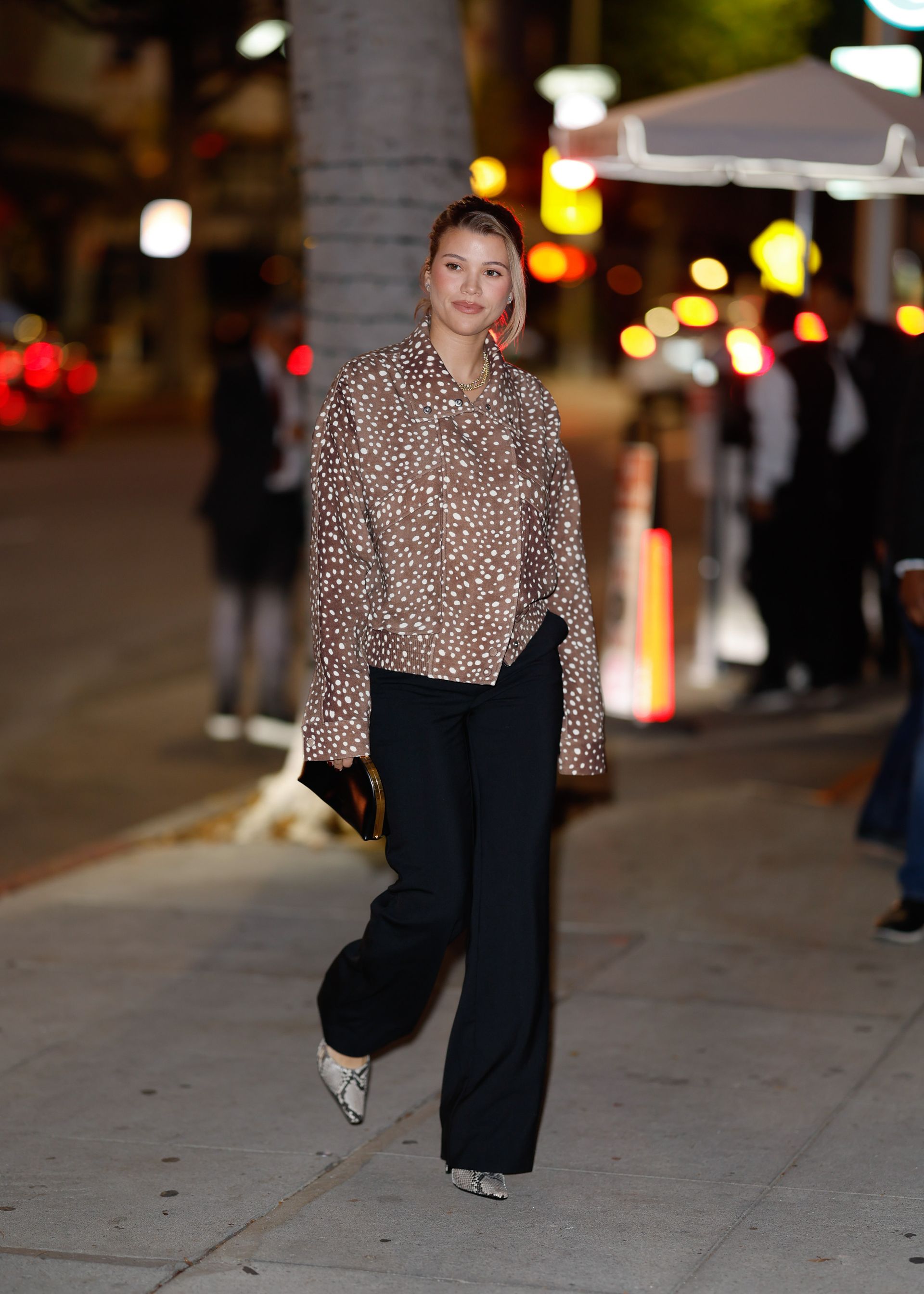 Sofia Richie walks down the street wearing black trousers with snakeskin heels and a deer print jacket.