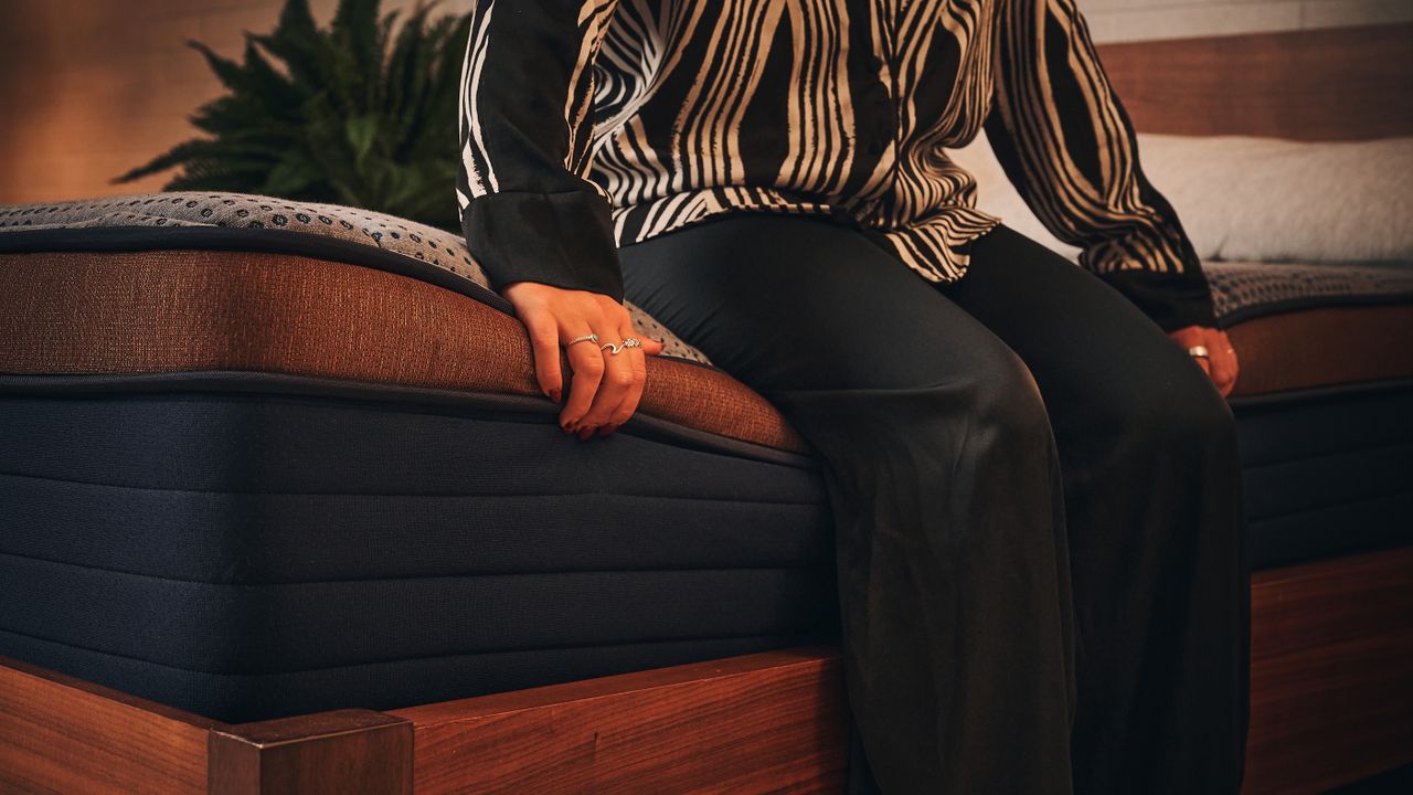 A woman in black and white silk pyjamas sitting on edge of Brooklyn Bedding Memory Foam Mattress on wooden bedframe in warmly lit sleep studio