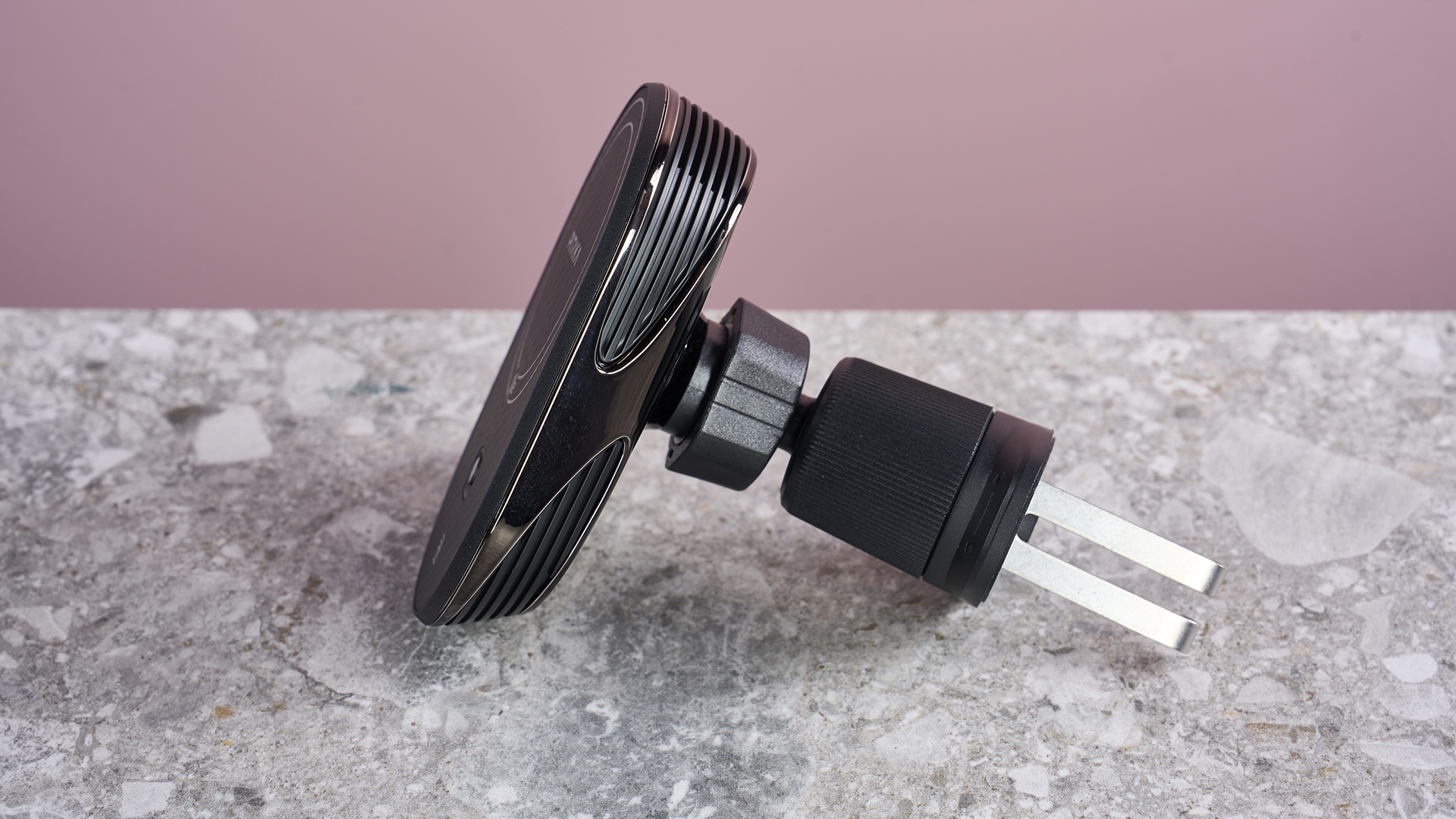 The Pitaka MagEZ Car Mount Pro 2 Qi2 sits on a stone-effect surface with a pink background. The view is of the side profile, revealing the collar fasteners and the metal attachment hooks.