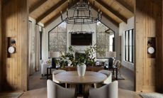 Open-concept rustic home interior featuring a round dining table in the foreground, leading into a high-ceilinged living room with large geometric windows and vaulted wood beams.