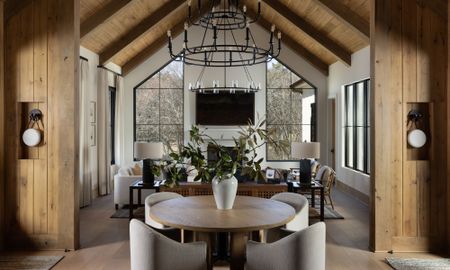 Open-concept rustic home interior featuring a round dining table in the foreground, leading into a high-ceilinged living room with large geometric windows and vaulted wood beams.