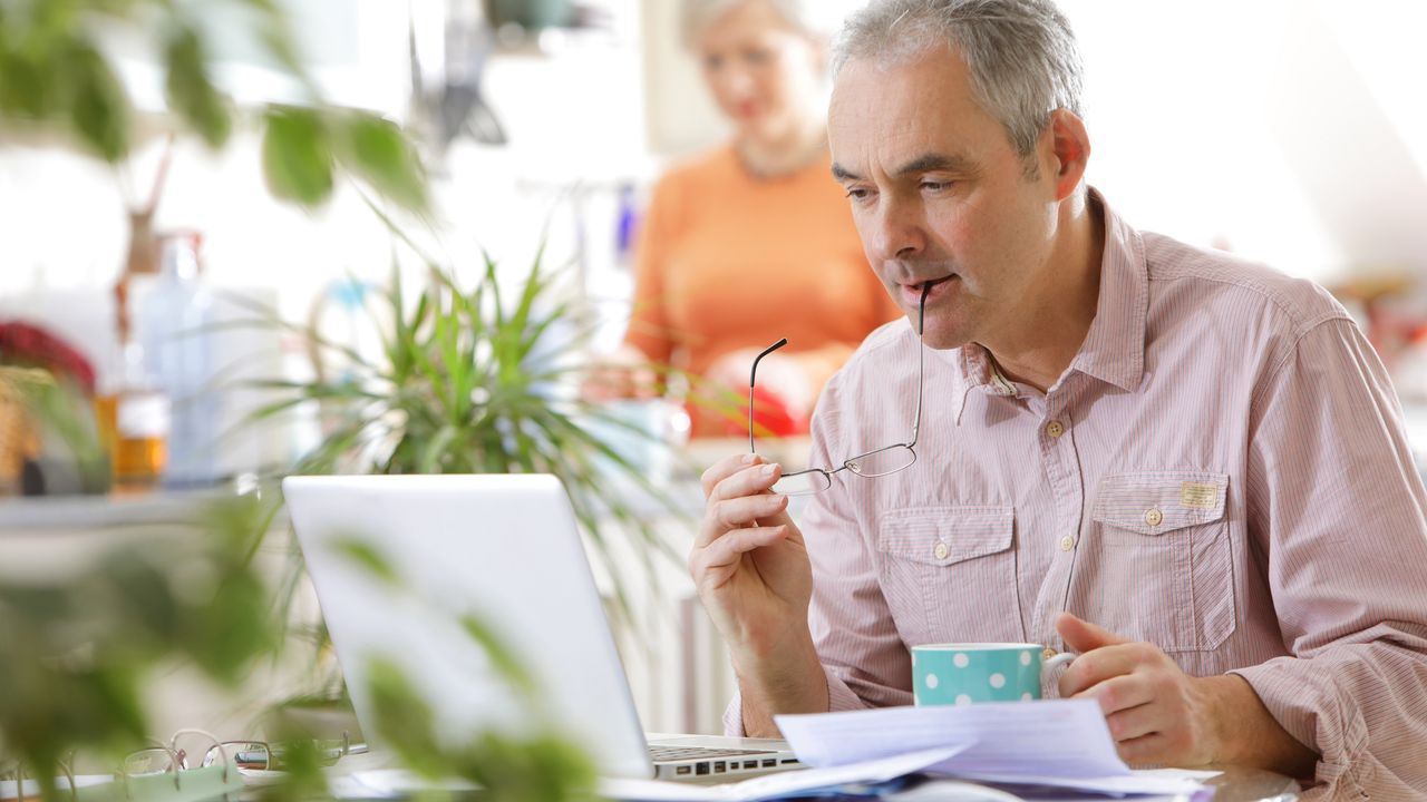 An older man looks at paperwork and his laptop at the kitchen table while his wife makes a cup of tea in the background.