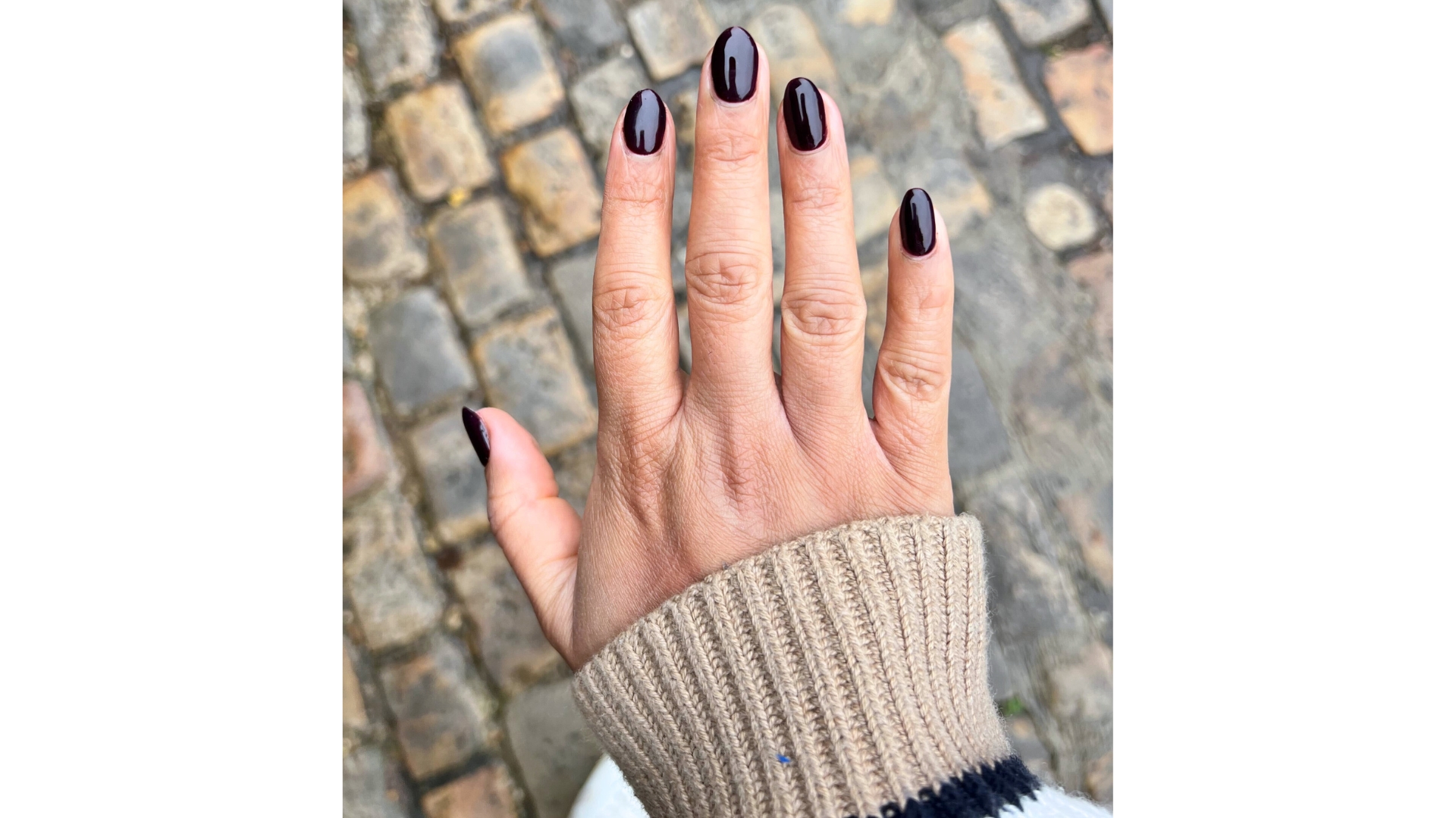 A close-up of Digital Beauty Ecom Editor Aleesha Badkar&#039;s hand, which features almond-shaped dark cherry nails/ pictured above a cobbled street