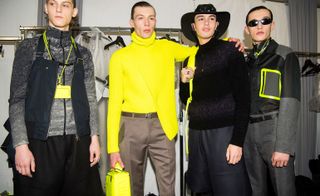 Four male models wearing looks from Louis Vuitton's collection. One model is wearing a grey jacket with blue sleeveless piece over the top, a yellow lanyard and black trousers. The model next to him is wearing a yellow asymmetric jumper, brown trousers, belt and is holding a yellow bag. The third model is wearing a black cowboy hat, black jumper with two lighter coloured stripes, dark grey trousers and is holding a yellow backpack. And the fourth model is wearing sunglasses, a grey, black and yellow shirt, necklace, belt and grey trousers