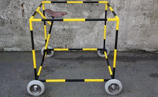 Concrete floor and wall, black and yellow striped metal frame, sat on four small grey, brown bicycle seat set inside the top of the frame