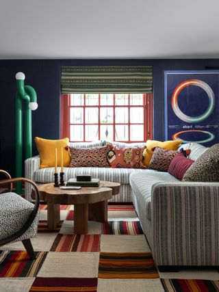 Image of a colorful living room. There is a striped sectional sofa with a wooden coffee table in front of it. The rug is a striped checkerboard pattern and the curtain on the window is a striped fabric as well. There is a green modern floor lamp beside the sofa.