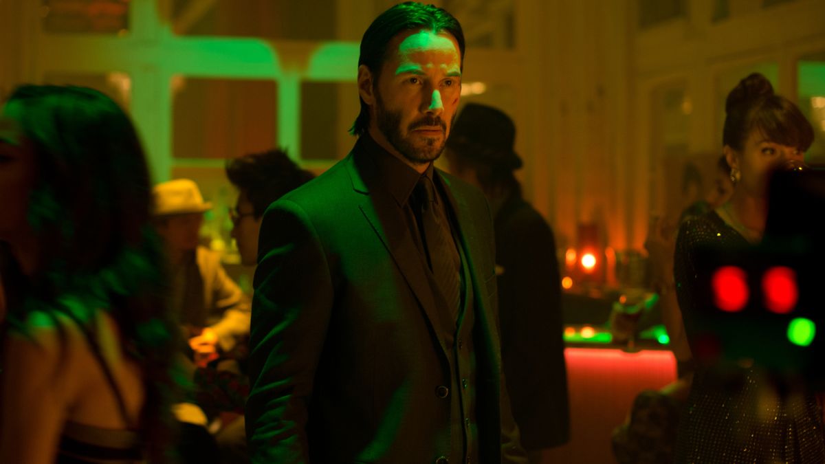 Keanu Reeves stands mysteriously in a crowded scene bathed in green light in John Wick.