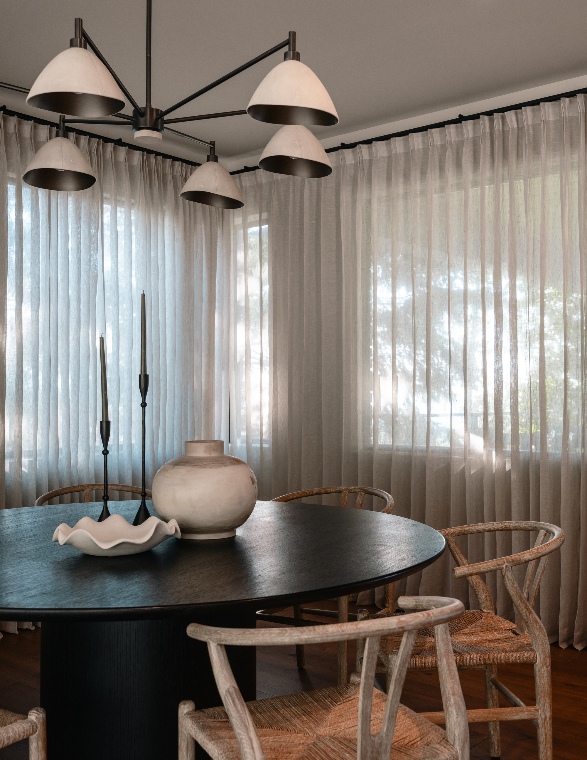 Image of a small dining room with a round, black dining table that has wooden chairs around it and a pendant light overhead. There are windows around the walls with sheer gray curtains over them.