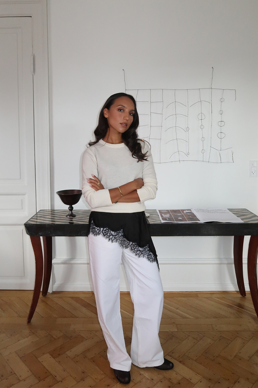 A sweater styling trend is shown in a photo of a woman&#039;s outfit, where she&#039;s standing inside her home in a hallway with a table and art behind her, wearing a white crewneck sweater over a black slip dress layered on top of white baggy jeans with black flats