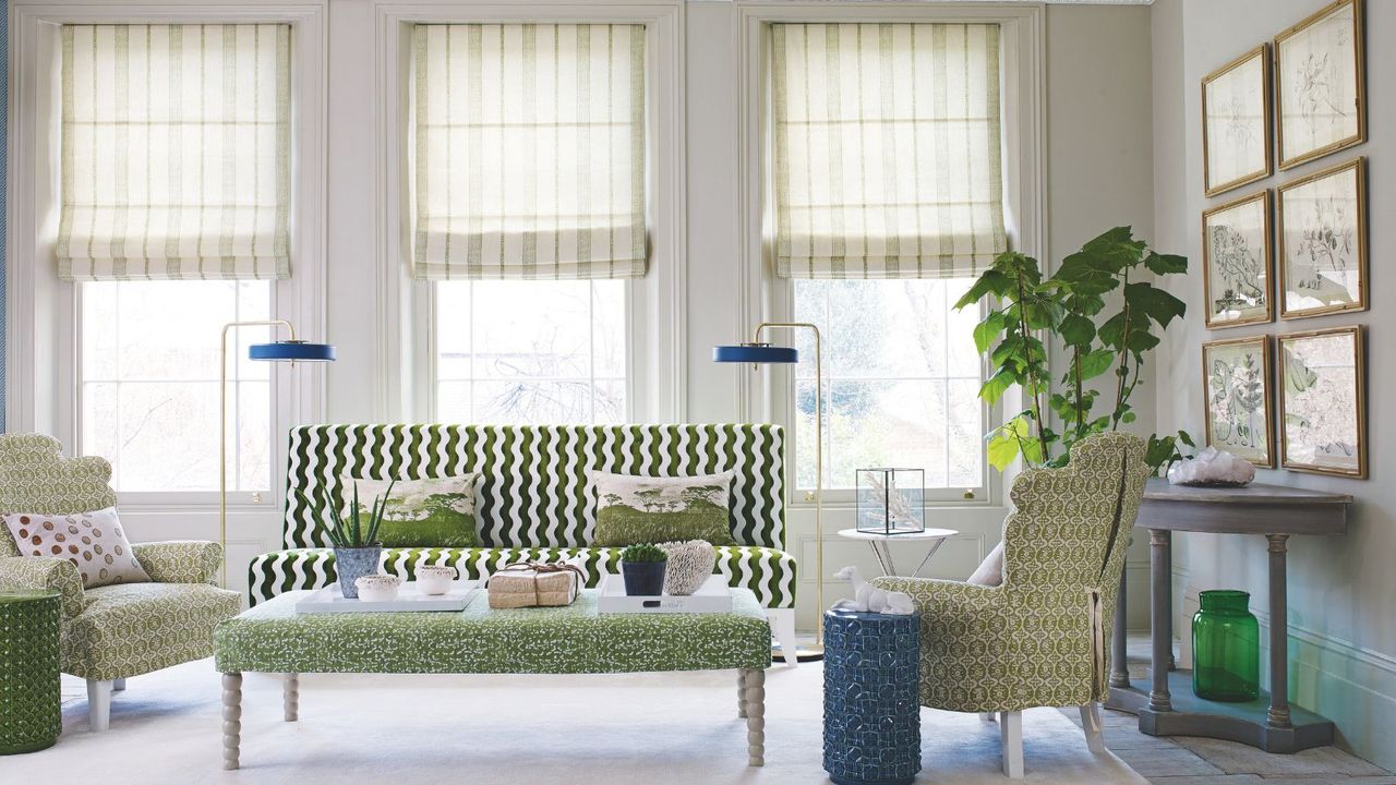 A bright living room with large windows with geometric blinds, and patterned geometric sofa, matching armchairs and padded footrest in green patterns, two free-standing gold and blue lamps, green and blue cylindrical coffee tables, a large plant and framed gallery wall above a grey sideboard