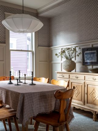 Silk pendant light hanging above a dining room table. The table has a tablecloth and five iron candlestick holders on it. There are rustic wooden chairs around the table