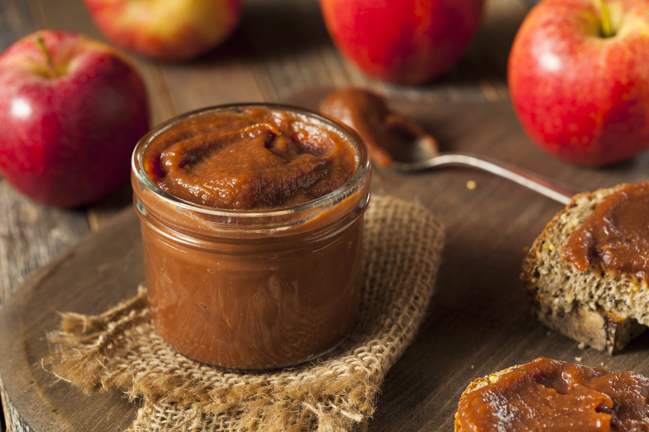 Homemade sweet apple butter with cinnamon and nutmeg in a jar on a table with a spoon, whole wheat bread, and apples.