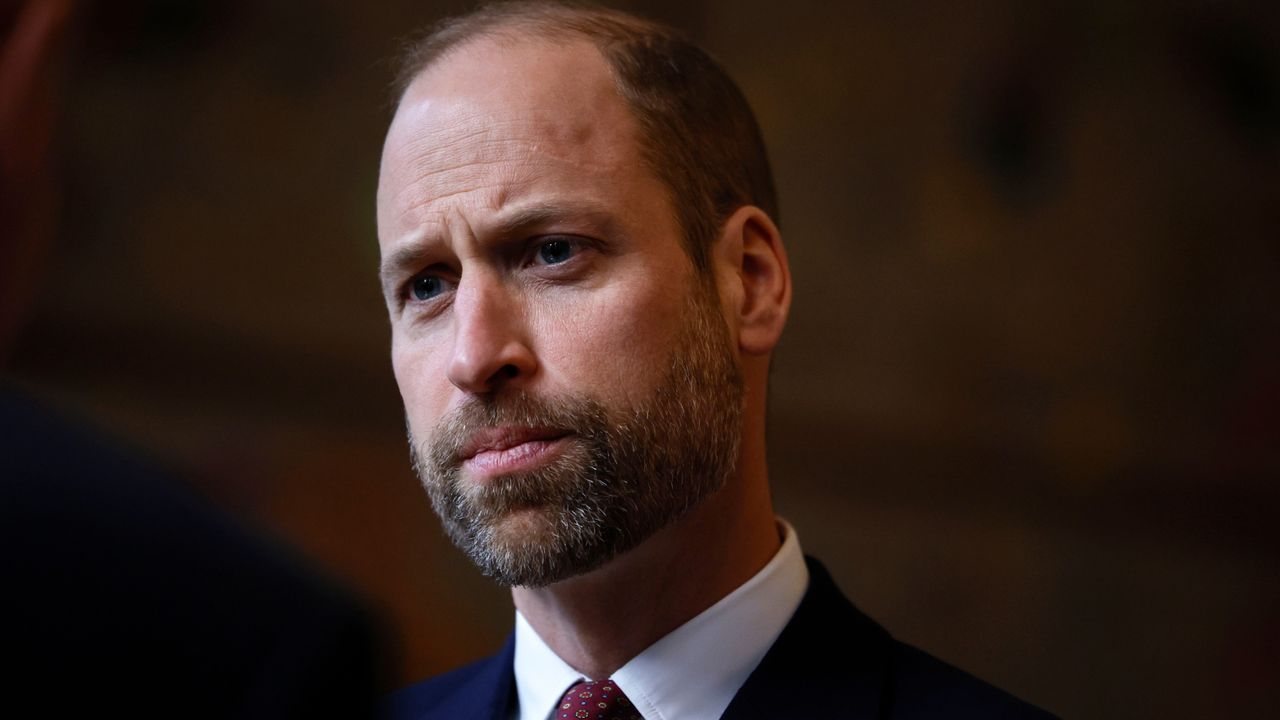 A shoulders-up photo of Prince William wearing a white shirt and red tie looking serious