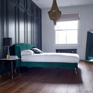 bedroom with blue panelled wall, hardwood floor, jade green bed, white bedding, gold chandelier, black and gold side table and table lamp, mirror