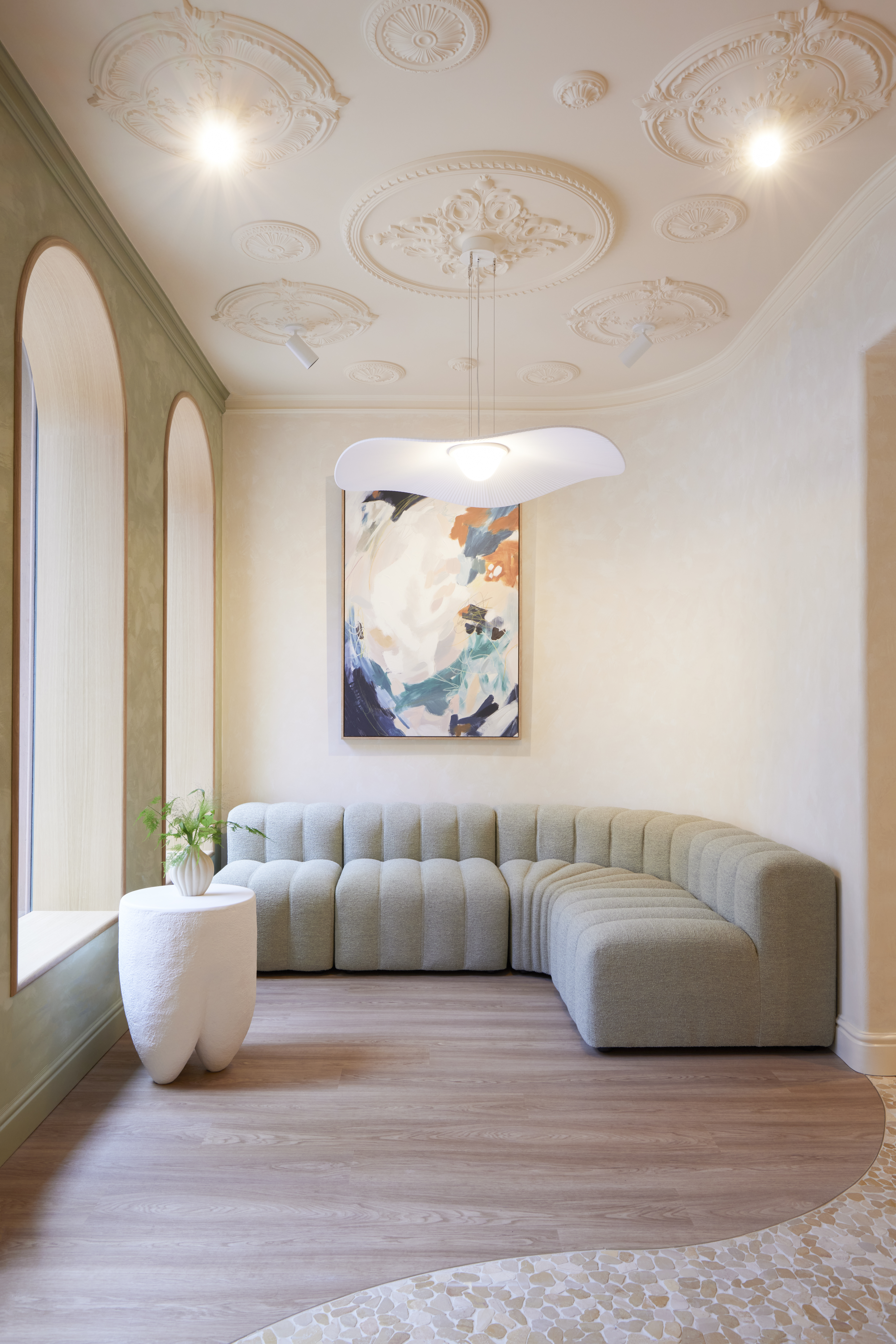 A pastel-shaded lobby boasts wooden flooring decorated with a wavy tiled mosaic, chunky ceramic furniture, a modular, water-green sofa, an organic pendant light shade, and arched windows.