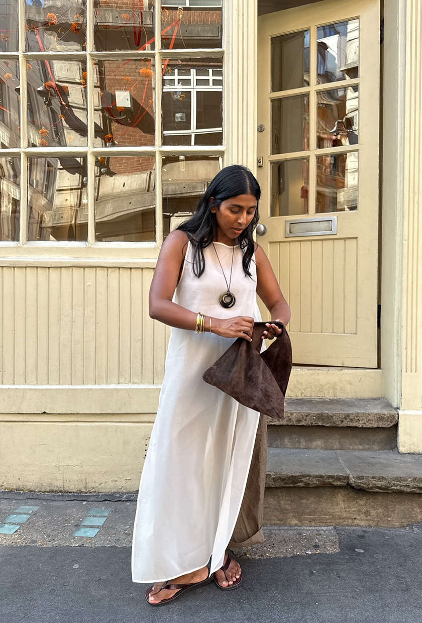 The coastal boho trend is shown in a street style photo of a woman standing outside in front of a coffee shop, wearing a white sheer maxi dress styled over olive green linen trousers worn with a long cord necklace, gold bangles, a dark brown suede hobo bag, and brown flip flops