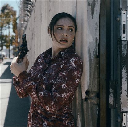 a woman (Juliana Aidén Martinez as June Hawkins) hides behind a wall while holding a gun