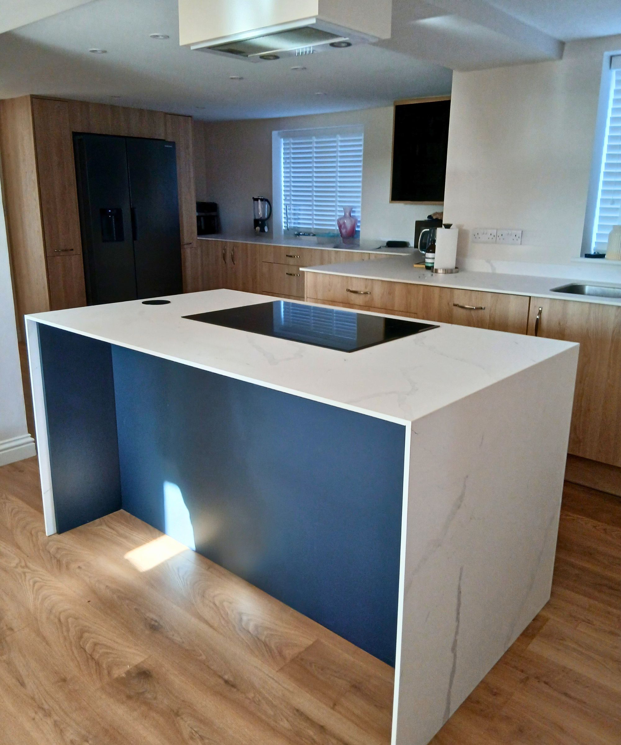 A modern kitchen with warm wood-effect flooring features a central island clad in white marble-look surfaces with subtle grey veining. The island has a deep navy inset panel and an integrated induction hob.
