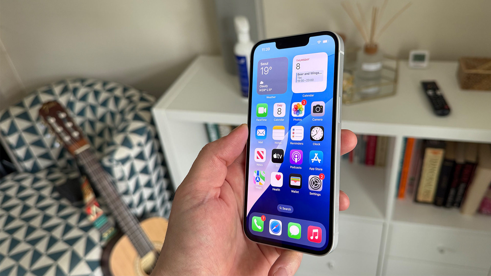 Apple iPhone 16e smartphone held in hand showing home screen with shelves and guitar in background