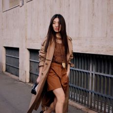 Woman wears toffee brown top and shorts with a tan trench coat while posing and walking for the camera