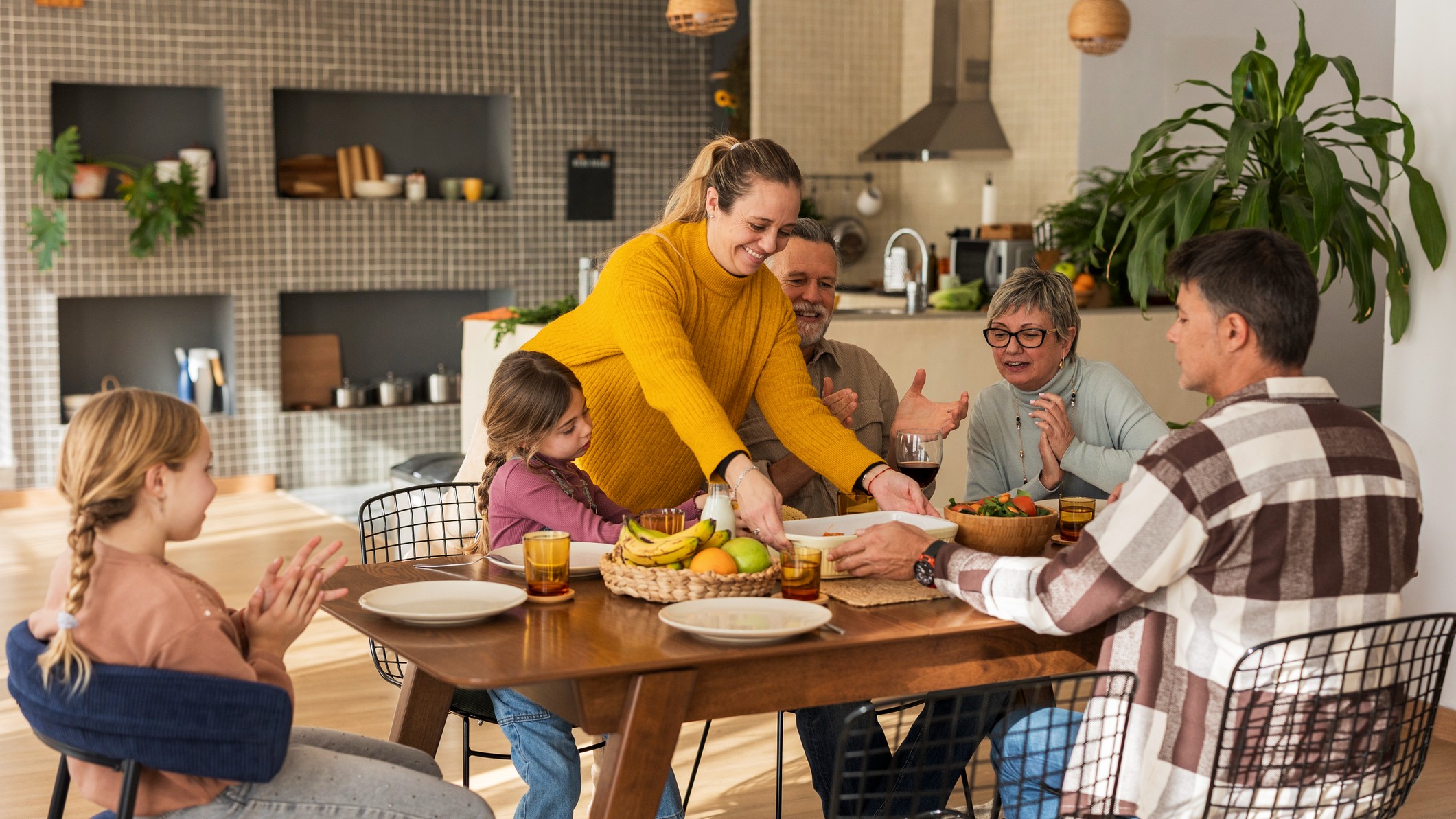 Three generations are living under one roof, or at least enjoying a meal together at home. The adult daughter is serving a dish while her spouse and two children look on. The grandparents are also at the table.