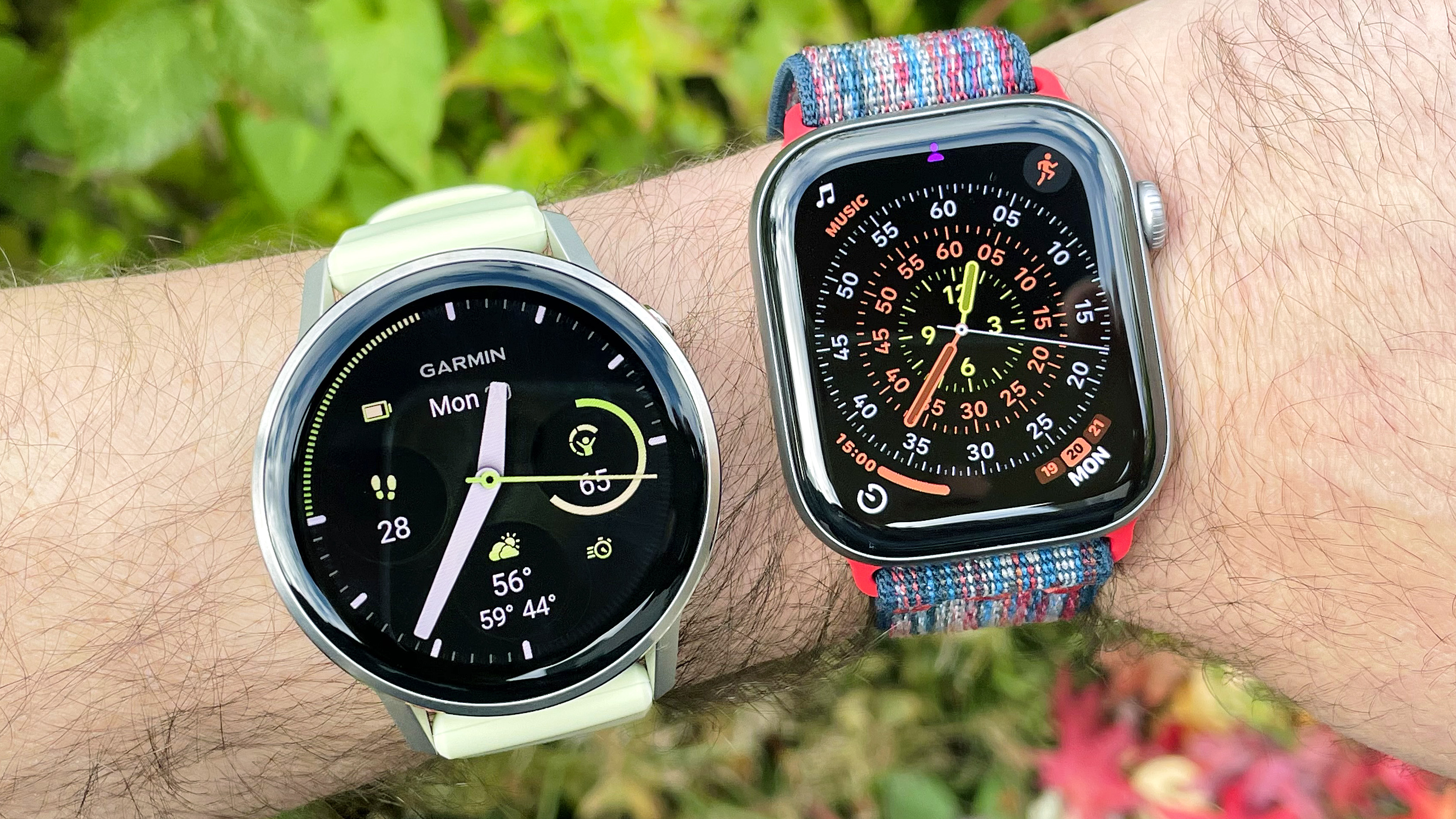 Close-up of the Garmin Venu 4 next to the Apple Watch Series 11 on a user's wrist with foliage out of focus in the background