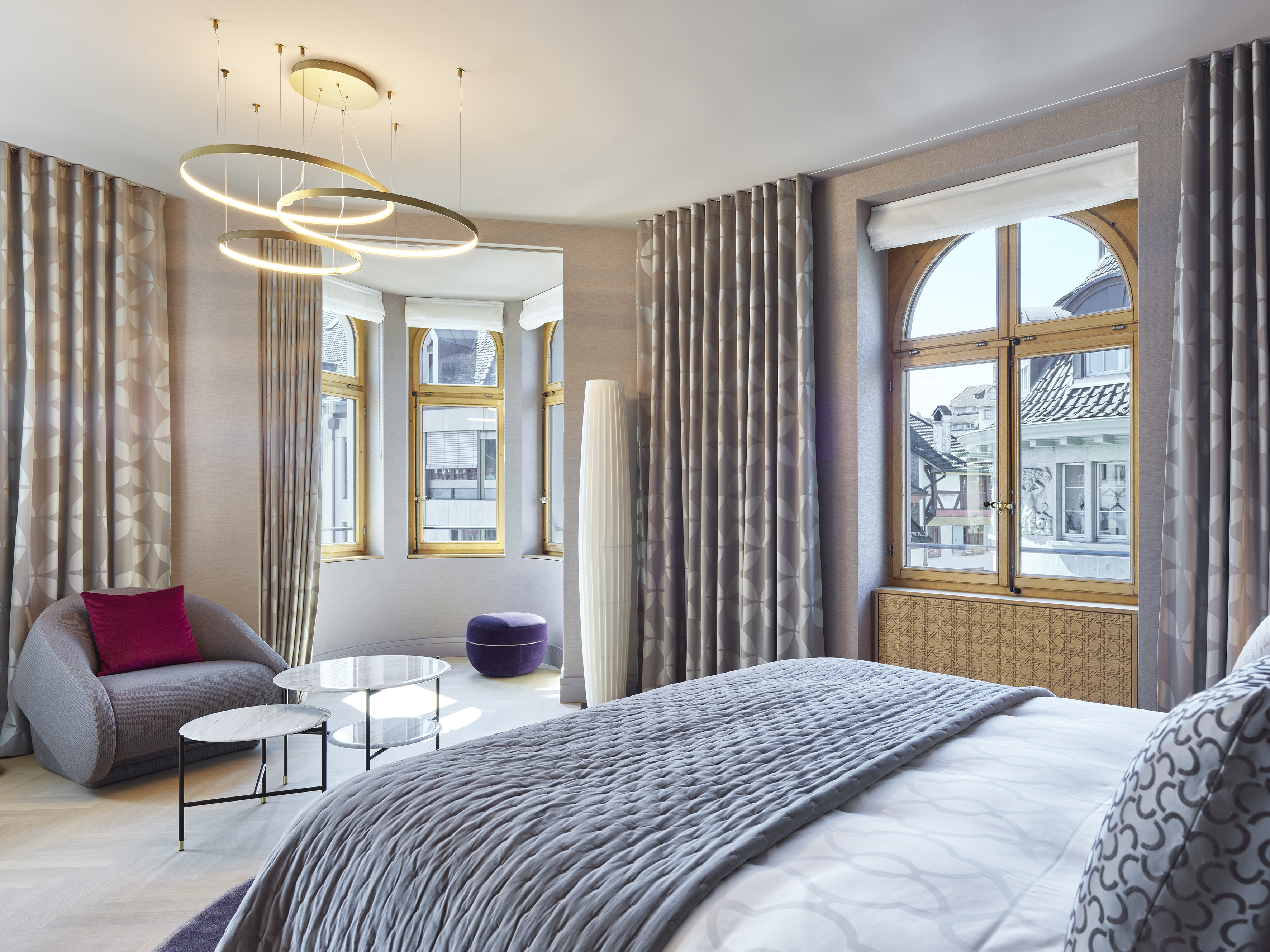 Photos of the interior of Hotel M&amp;auml;rthof Basel shows a gray blanket covering a white bedspread, with retro style furniture and large arched windows.