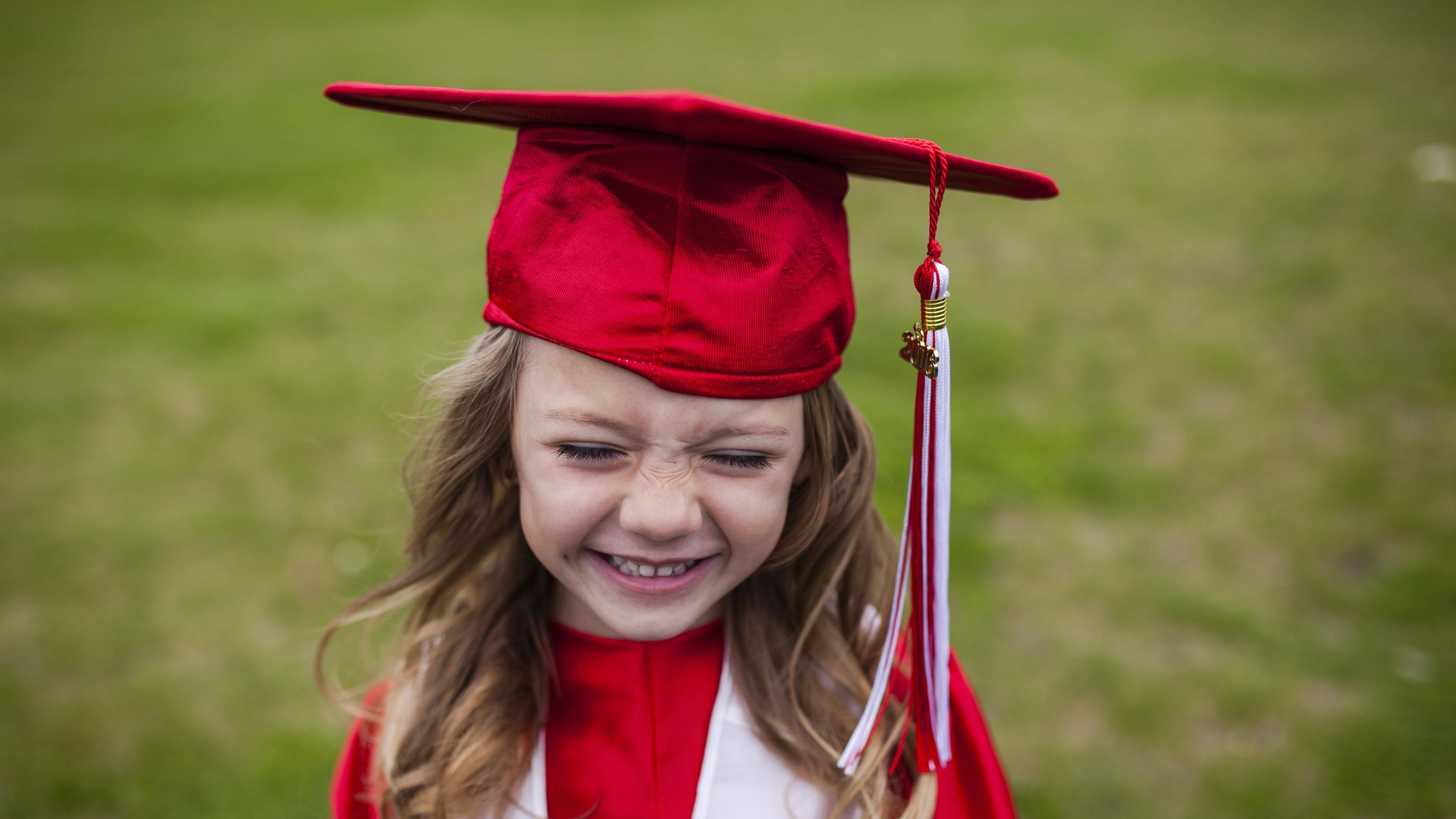 A cute little girl smiles while wearing red cap and gown and standing in a field.