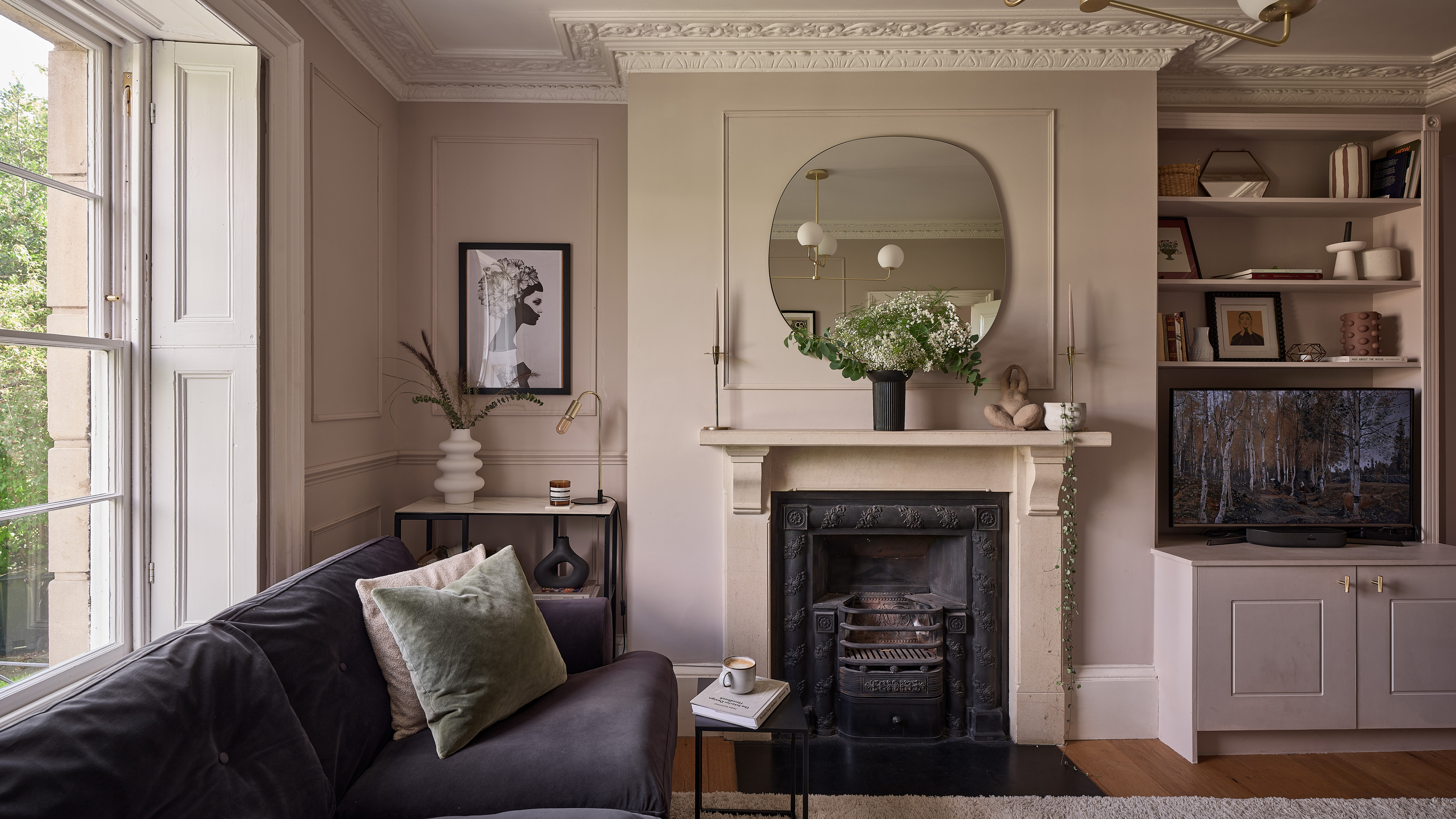 a pale pink living room with a dark purple velvet sofa fireplace, large circular overmantel mirror and classic cornicing