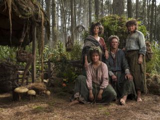 (L to R) Markella Kavenagh (as Elanor ‘Nori’ Brandyfoot), Sara Zwangobani (as Marigold Brandyfoot), Dylan Smith (as Largo Brandyfoot), Megan Richards (as Poppy Proudfellow) sit in front of a hut in The Lord of the Rings: The Rings of Power
