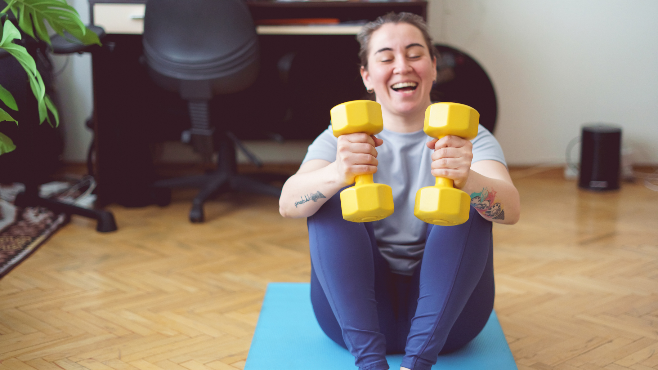 Woman holding dumbbells while sitting on an exercise mat on a wooden floor in a domestic setting. She has her eyes closed, mouth open and is smiling