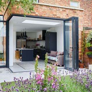 brick wall house with a glass extension with retracting doors and an indoor brown sofa with cushions and a colorful throw next to a black wooden kitchen