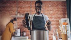 Man wearing an apron about to stir the contents of a pressure cooker with a wooden spatula