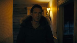 A man is wearing a thick winter jacket as he searches a dimly light wooden cabin at night.