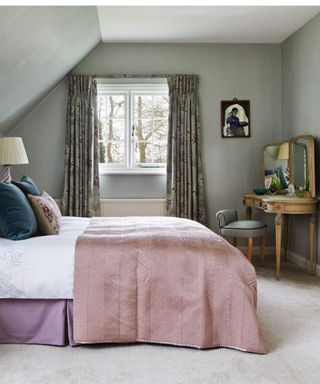 Peaceful bedroom with sloped ceiling, plush cream carpets and ornate gold dressing table with three-part mirror. There is a door open and a sliver of an en suite bathroom visible