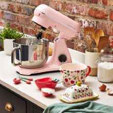 A pink Lakeland mixer on a kitchen counter with other kitchen utensilswith 