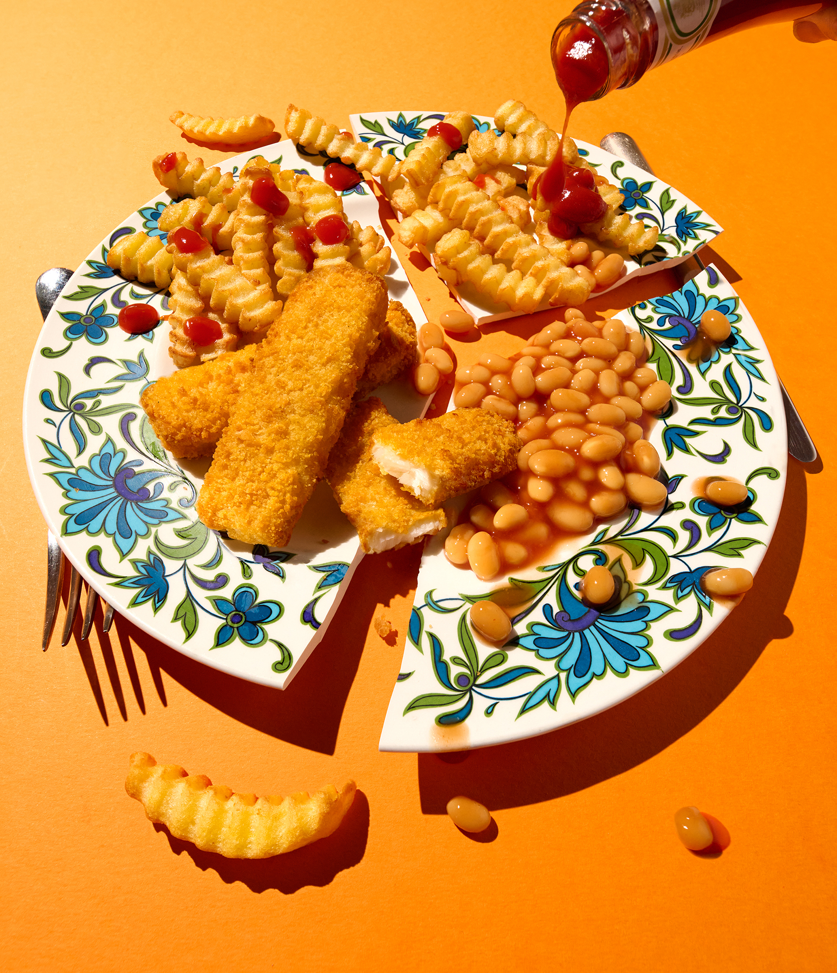 A broken plate holds crispy fish fingers, baked beans, and crinkle fries drizzled with ketchup against a bright orange background